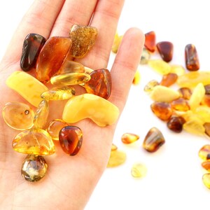 May include: A collection of amber stones in various shades of yellow, orange, and brown. The stones are smooth and polished, and they are arranged in a pile on a white surface.