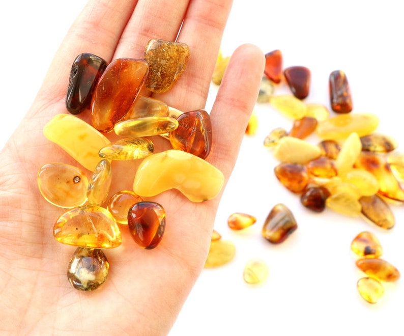 May include: A collection of amber stones in various shades of yellow, orange, and brown. The stones are smooth and polished, and they are arranged in a pile on a white surface.