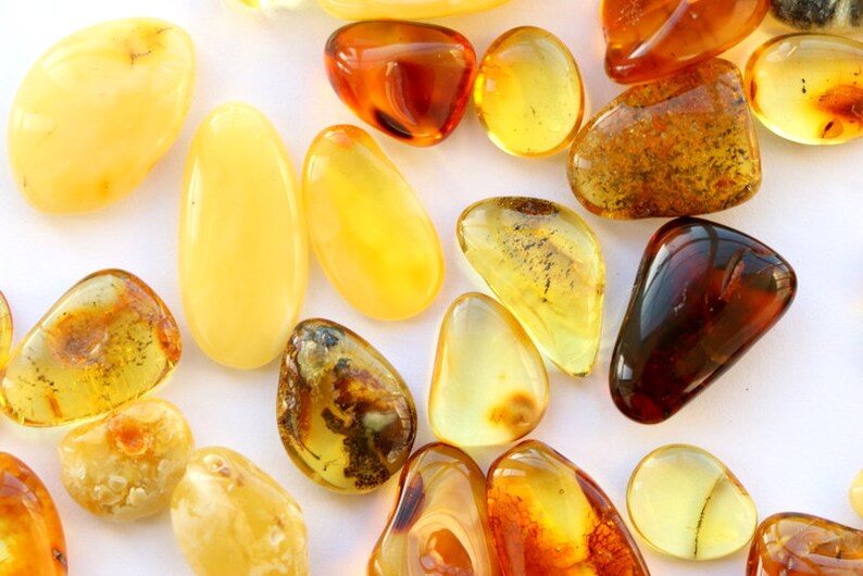 May include: A collection of amber stones in various shades of yellow, orange, and brown. The stones are smooth and polished, with some showing inclusions of insects or plant matter.