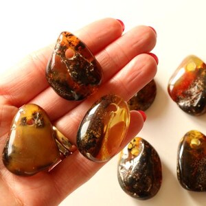 May include: A collection of seven amber gemstone pendants, each with a unique shape and color variation. The pendants are a mix of brown, orange, and yellow hues, with some featuring inclusions of natural materials.