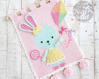 May include: A handmade crochet wall hanging featuring a light blue bunny holding a lollipop and a kite. The pink banner has a scalloped edge and pom-poms. The bunny wears a yellow dress and pink shoes. The banner is suspended from a wooden dowel.