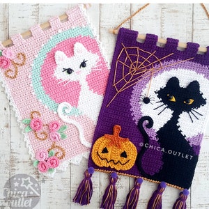 Puede incluir: Dos tapices de pared de ganchillo con motivos de gatos. El primero es rosa con un gato blanco y un diseño floral. El segundo es morado con un gato negro, una telaraña y una calabaza. Ambos tapices tienen listones de madera en la parte superior y borlas en la parte inferior.  @CHICA.OUTLET