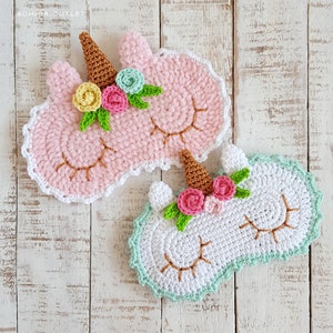 May include: Two handmade crochet unicorn sleep masks. One is pink with a white trim, and the other is white with a mint green trim. Both have a gold horn, closed eyes, and floral accents in shades of pink, yellow, and green.