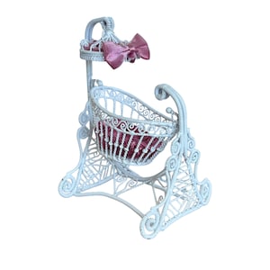 May include: A white wicker baby cradle with a pink bow and a pink interior. The cradle has a decorative design and is made of woven wicker. The cradle is designed to rock gently. A charming nursery decor item.