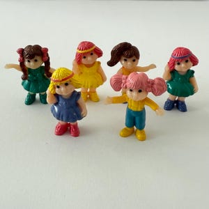 Pode incluir: Seis pequenas bonecas de plástico com vários penteados e roupas coloridas. As bonecas têm vestidos, tops e calças de cores diferentes. As bonecas são em tons de verde, amarelo, azul e rosa. As bonecas estão em uma superfície branca.