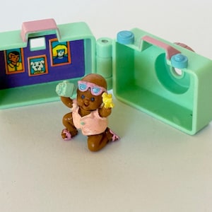 Pode incluir: Um conjunto de brinquedos vintage com uma boneca marrom em miniatura com óculos de sol e uma câmera, ajoelhada em frente a uma caixa em forma de câmera verde-menta. A caixa tem uma alça rosa e detalhes em azul. A boneca está segurando uma flor amarela.