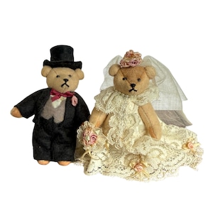 May include: Two teddy bears dressed for a wedding. One bear wears a black tuxedo with a top hat and a pink bow tie. The other bear is in a cream-colored lace wedding dress, holding a bouquet of pink roses and wearing a veil.