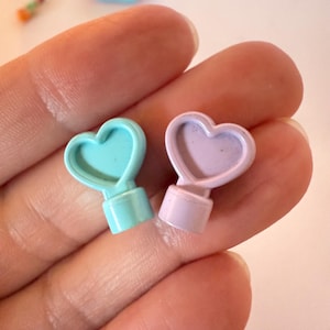 Pode incluir: Dois pequenos objetos em forma de coração em cores pastel. Um é azul claro, o outro é lavanda. Cada um tem uma base cilíndrica e um topo em forma de coração. Os objetos são segurados em uma mão.