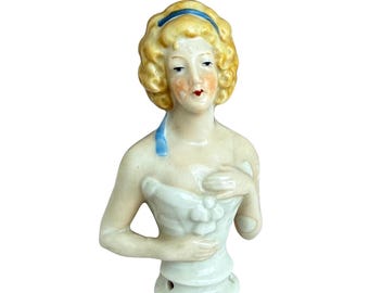 May include: A vintage porcelain half-doll figurine with blonde curly hair and a blue headband. The doll has a delicate face with painted features and is wearing a white bodice. The figurine is a collectible item.