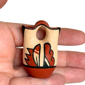 May include: A small, hand-painted ceramic vase with a handle and a hole. The vase features a cream-colored body with red and black geometric designs. The base and rim are painted red.