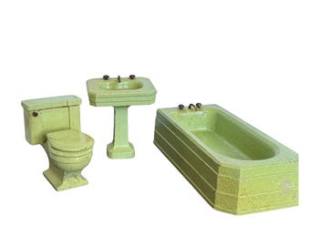 Puede incluir: Un juego de baño en miniatura de estilo vintage, que incluye un inodoro, un lavabo con pedestal y una bañera. El conjunto está pintado en un color verde descolorido, con desgaste visible, lo que sugiere una estética antigua. Los grifos tienen detalles de color latón.