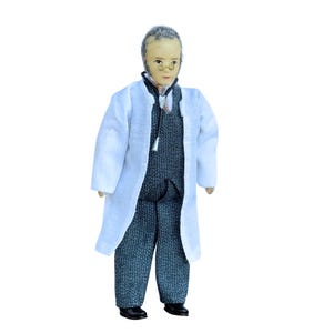 Puede incluir: Una muñeca en miniatura que representa a un médico, vestido con una bata de laboratorio blanca sobre un chaleco y pantalones grises. La muñeca tiene una cara pintada, gafas y pelo gris. Zapatos negros completan el atuendo. La muñeca está de pie.