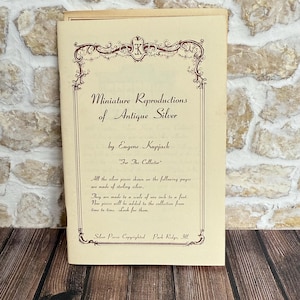 May include: An open booklet titled "Miniature Reproductions of Antique Silver" by Eugene Kupjack. The cover features ornate brown decorative borders and text. The text states the silver pieces are made of sterling silver and are scaled at one inch to a foot.