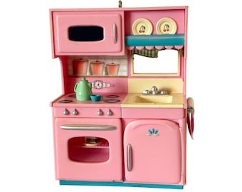 May include: A pink toy kitchen playset featuring a microwave, stove with four burners, oven, and sink. The unit includes a light green kettle, a small blue cup, two plates with cherry illustrations, and a towel hanging on the side handle.