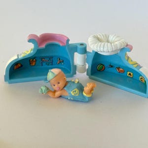 Pode incluir: Figura vintage de bebê com roupa e boné azuis, em frente a um recipiente em forma de sapato azul e branco aberto. O interior do recipiente é rosa e branco, com pequenas ilustrações. Um objeto de coleção charmoso.