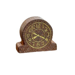 Op de afbeelding: Een kleine houten klok met een bruine afwerking en goudkleurige cijfers en wijzers. De klok heeft een afgeronde bovenkant en een rechthoekige basis. Hij is ontworpen om op een oppervlak te staan. De klok is ongeveer 5 cm hoog.