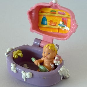 Pode incluir: Um conjunto de brinquedos em miniatura vintage com uma boneca em uma banheira lilás com bolhas de sabão. O conjunto inclui uma prateleira rosa com uma ilustração de pato e garrafa. A boneca segura uma escova verde e uma esponja rosa.
