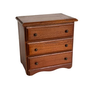 May include: A miniature wooden dresser with three drawers and dark knobs. The dresser is a warm brown color and has a slightly textured surface. The top of the dresser has a small overhang.