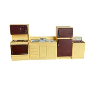 May include: A miniature kitchen set in shades of yellow and brown. The set includes a stove with a microwave, a sink with a countertop, a refrigerator, and a washing machine. The appliances have brown doors and yellow frames.