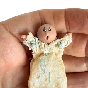 May include: A miniature doll with a round, light-toned head, dark eyes, and a small mouth. The doll wears a cream-colored fabric gown featuring a lace collar and light blue ribbon accents at the sleeves and center, with arms extended outward.