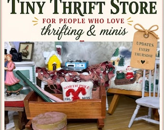 May include: A miniature scene featuring a wooden cradle, a small white pillow with red North Pole embroidery, a yellow house figurine, a blue stove, and a tag reading UPDATES EVERY THURSDAY. The text reads VINTAGE DOLLHOUSE SHOP TINY THRIFT STORE.
