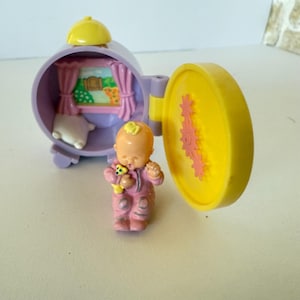 Pode incluir: Um conjunto de brinquedos vintage com uma figura de bebê em um macacão rosa segurando um ursinho de pelúcia amarelo. O conjunto inclui uma caixa circular roxa com uma porta amarela, uma janela com cortinas e uma almofada.