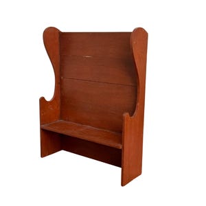 May include: A small, antique-style wooden bench painted in a reddish-brown hue. The bench features a high, curved back and a simple seat. The design is reminiscent of a classic, rustic aesthetic.