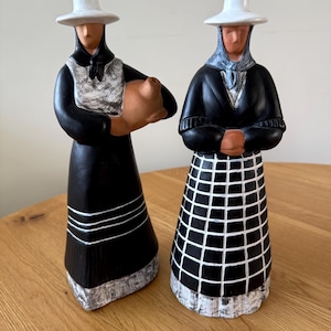 May include: Two ceramic figurines depicting women in traditional dress. One holds a brown jug, the other has folded arms. Both wear black dresses, white hats, and have white and grey accents. The figurines are on a wooden surface.
