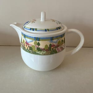 May include: White ceramic teapot with a lid. The teapot features a colourful landscape design with houses, trees, and fields. The lid has a floral pattern and a handle. The teapot has a curved handle and a spout.