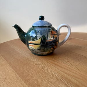 May include: A ceramic teapot with a painted coastal scene featuring boats and buildings. The teapot has a dark green spout and handle, a light blue lid, and a dark green knob. The teapot is sitting on a wooden surface.