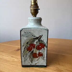 May include: A rectangular ceramic lamp base with a speckled off-white glaze. The front features a hand-painted botanical illustration of red berries and green leaves. A brass-coloured metal light socket is attached to the top of the ceramic body.