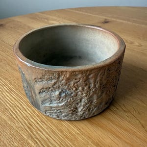 May include: A handmade, round ceramic bowl with a textured exterior. The bowl has a natural colour palette with shades of brown, tan, and grey. The interior is a darker shade of grey. The bowl sits on a wooden surface.