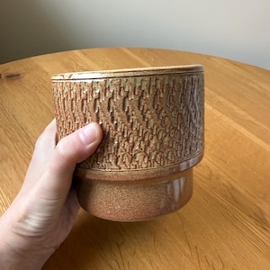 May include: A brown ceramic plant pot with a textured, bark-like pattern. The pot has a cylindrical shape with a slightly flared base. The colour is a warm, earthy tone, suitable for indoor or outdoor use.