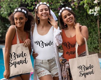 Op de afbeelding: Drie vriendinnen dragen witte bloemenkransen en houden tote bags vast met de tekst "Bride Squad" erop gedrukt. De tassen zijn gemaakt van natuurlijk canvas en hebben zwarte handvatten.
