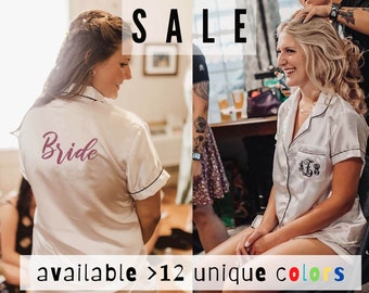 May include: Two women wearing white satin robes with embroidery. One robe says "Bride" in pink lettering and the other has initials. The text "SALE" is at the top of the image and "available > 12 unique colours" is at the bottom.