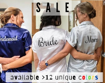 Puede incluir: Tres mujeres con batas de satén a juego con texto bordado. Las batas son azul marino, blanco y plateado. El texto en las batas dice "Bride", "Bridesmaid" y "Mom". El texto "SALE" está en la parte superior de la imagen. El texto "available > 12 unique colors" está en la parte inferior de la imagen.