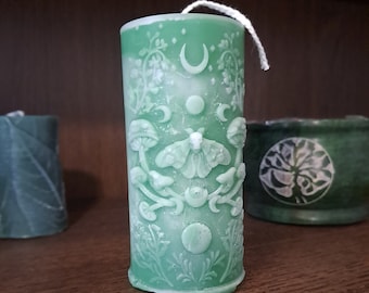 May include: A green pillar candle with a white wick and intricate carvings of moons, flowers, mushrooms, and a moth. The candle has a textured surface and is decorated with a white and green color scheme.