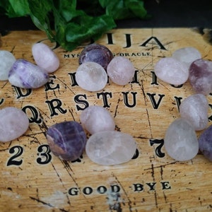 Könnte beinhalten: Eine Nahaufnahme eines Ouija-Bretts mit einer Vielzahl kleiner, getrommelter Amethyst- und Rosenquarzkristalle. Die Kristalle sind auf dem Brett angeordnet, wobei einige von ihnen auf den Buchstaben und Zahlen liegen.