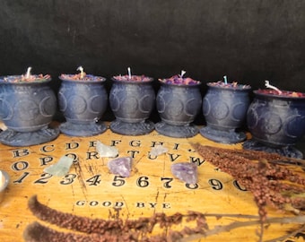 May include: Six black cauldron-shaped candles with purple and blue accents, arranged on a wooden surface with a Ouija board and crystals.