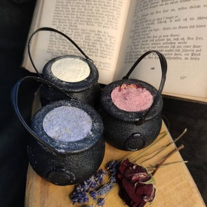 Könnte beinhalten: Drei schwarze, kesselartige Badebomben mit verschiedenen farbigen Badesalzen im Inneren. Die Badebomben befinden sich auf einer Holzoberfläche mit getrockneten Lavendel- und Rosenblüten.