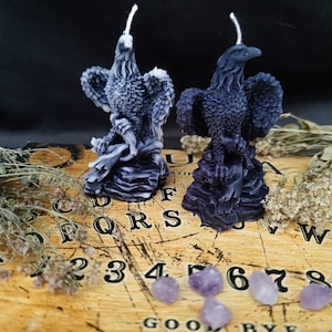 Könnte beinhalten: Zwei schwarze und weiße Adlerkerzen auf einem Ouija-Brett aus Holz mit violetten Amethyst-Kristallen. Die Kerzen sind detailliert und haben eine strukturierte Oberfläche.