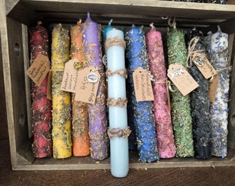 May include: A collection of 10 colorful candles with various herbs and crystals embedded in them. The candles are arranged in a wooden crate. The candles are labeled with names like "Freie Intuition", "Ausdauer", "Schutz", "Freundschaft", "Kraft & Spiritualle", "Schutz", "Kraft", "Schutz", "Schutz", and "Schutz".