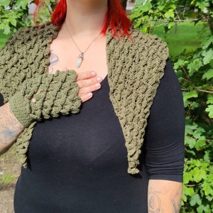 Könnte beinhalten: Olivgrüner Häkel-Schulterwärmer und passende fingerlose Handschuhe. Das strukturierte Strickwerk hat ein einzigartiges Muster und wird über einem schwarzen Oberteil getragen. Ein stilvolles Accessoire für kühlere Tage.
