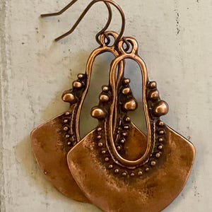 May include: A pair of copper earrings with a unique, ornate design. The earrings feature a large, fan-shaped pendant with a textured surface and a smaller, oval-shaped loop at the top.