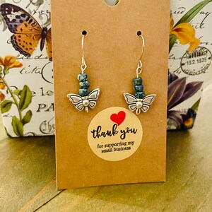 May include: A pair of silver butterfly earrings with blue and green gemstone beads. The earrings are displayed on a brown card with a thank you sticker.