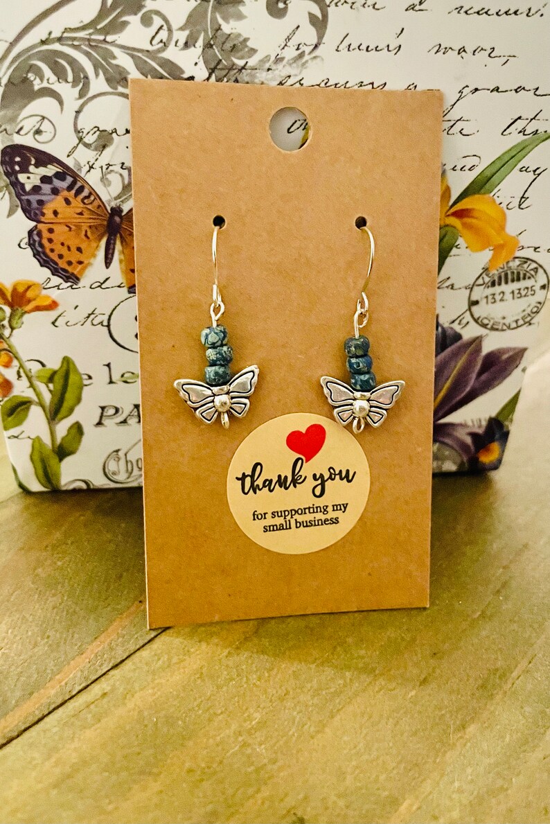May include: A pair of silver butterfly earrings with blue and green gemstone beads. The earrings are displayed on a brown card with a thank you sticker.