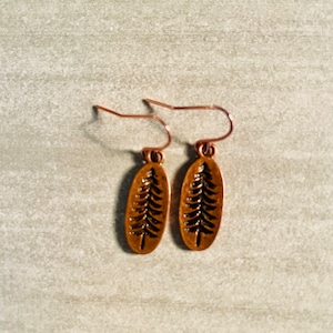 May include: A pair of copper earrings with a tree design. The earrings are oval shaped and have a wire hook.