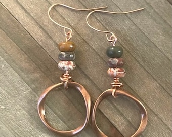 May include: A pair of dangle earrings featuring copper-toned wire hooks and circular frames. Each earring is accented with three stacked beads in shades of brown, gray, and mottled white, connected to the copper circle by wrapped wire detailing.