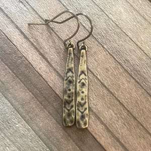 May include: A pair of bronze-colored earrings with a textured, elongated design. The earrings have a hook closure and feature a series of small, dark markings. The earrings are displayed on a wooden surface.