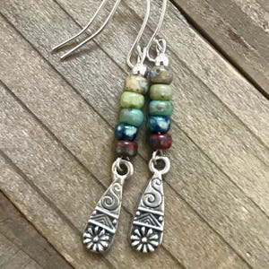 May include: A pair of dangle earrings with silver-tone hooks. Each earring features a vertical row of colorful beads in shades of green, blue, and red, topped with a silver-tone accent. The bottom of each earring has a silver-tone teardrop shape with a floral design.